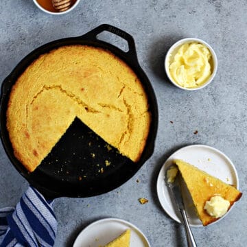 Looking down on cornbread baked in a cast iron skillet with two slices cut out and placed on small plates topped with butter and honey.