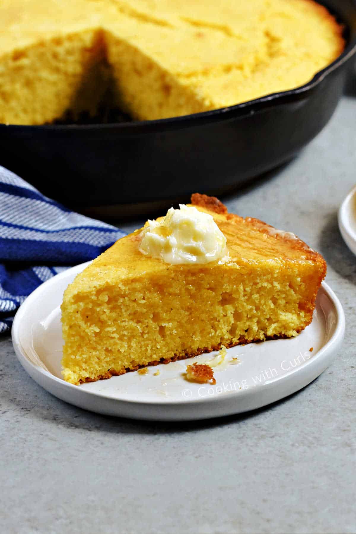 A wedge of skillet cornbread on a small plate topped with butter and honey with the skillet in the background.