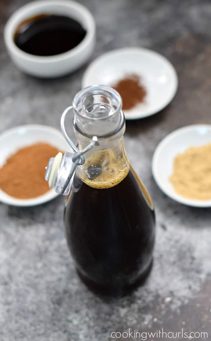 Clear bottle filled with gingersnap syrup with small white bowls of spices in the background.