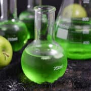 Three Poisoned Apple Cocktails in glass science bottles, two green apples, and a large beaker.