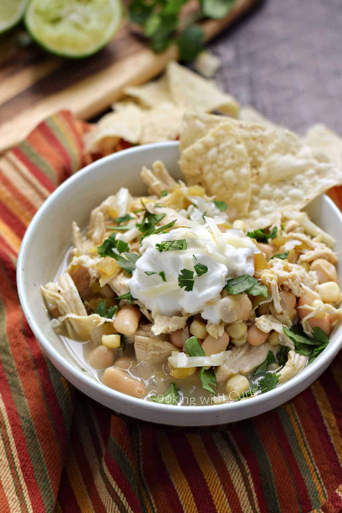 A bowl of white chicken chili topped with sour cream, cilantro, and tortilla chips with limes in the background.