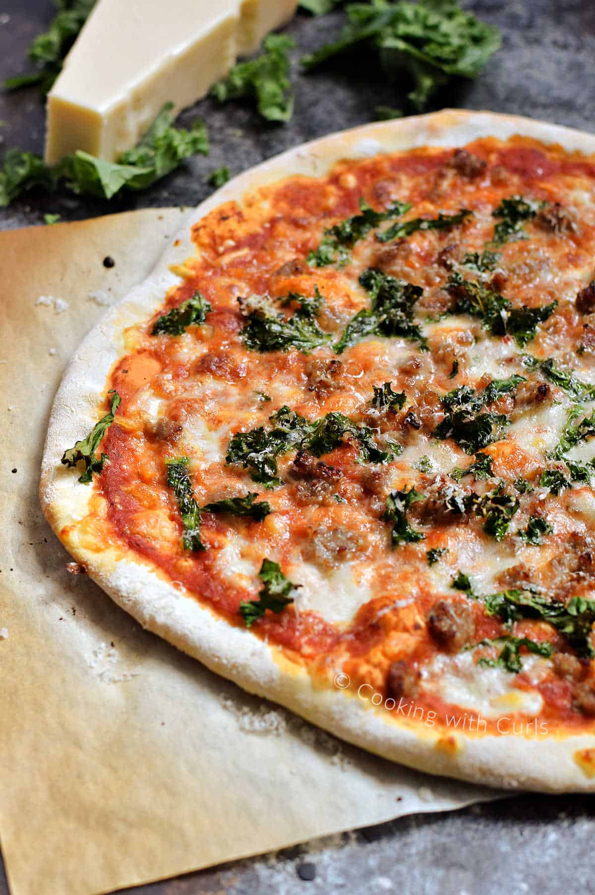 A whole pizza on parchment paper topped with tomato sauce, melted cheese, Italian sausage, and chopped kale with a wedge of parmesan in the background. 