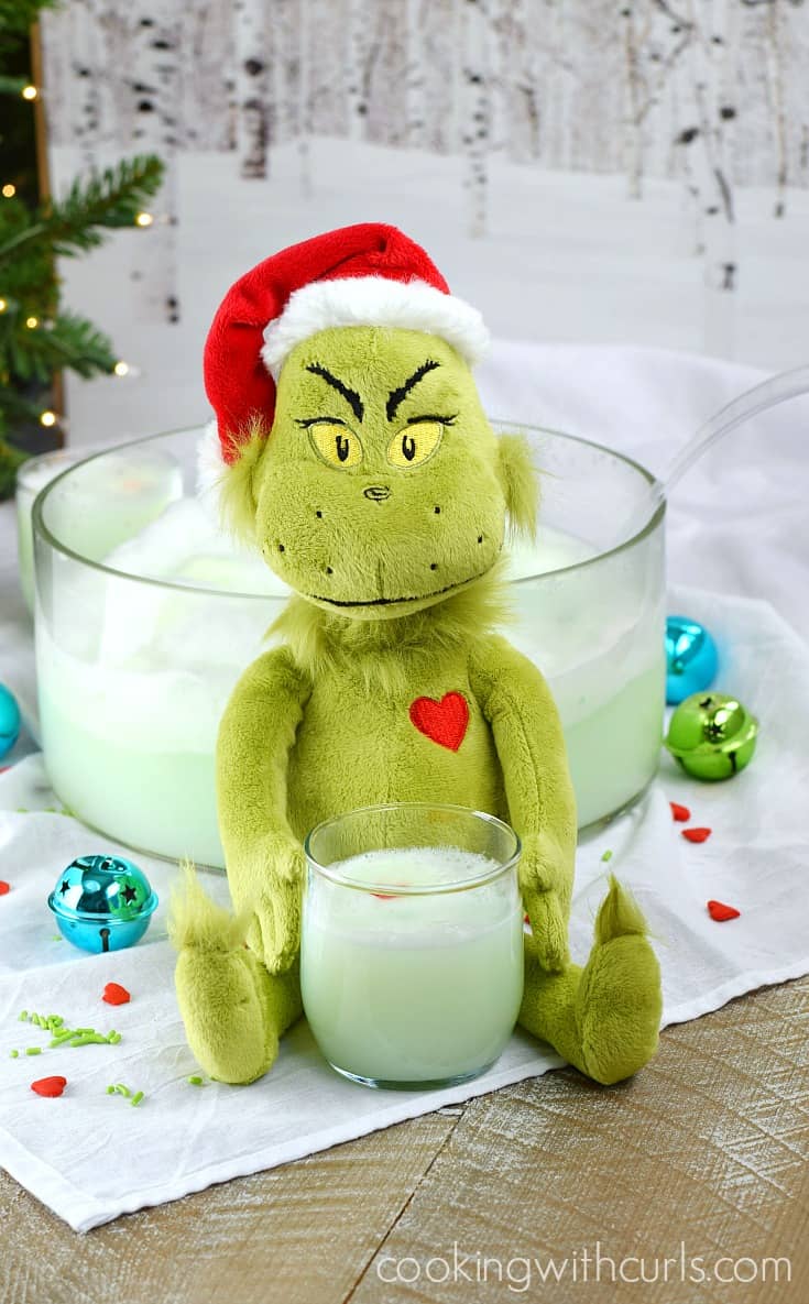 A stuffed grinch doll sitting in front of a punch bowl filled with green punch with a mug of punch in between his hands.