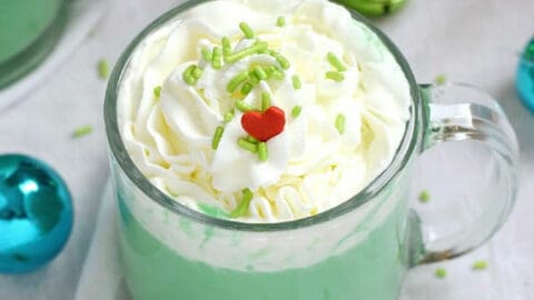 A glass mug filled with green hot chocolate topped with whipped cream, green sprinkles, and a red heart.