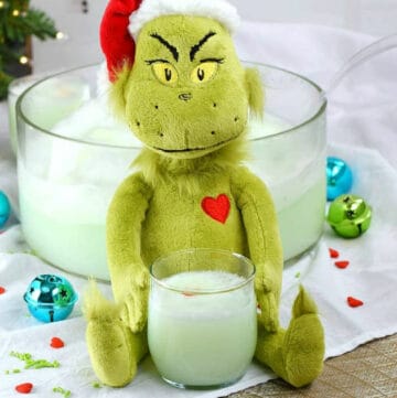 A punch bowl filled with green sherbet punch behind a stuffed Grinch toy holding a glass of punch.