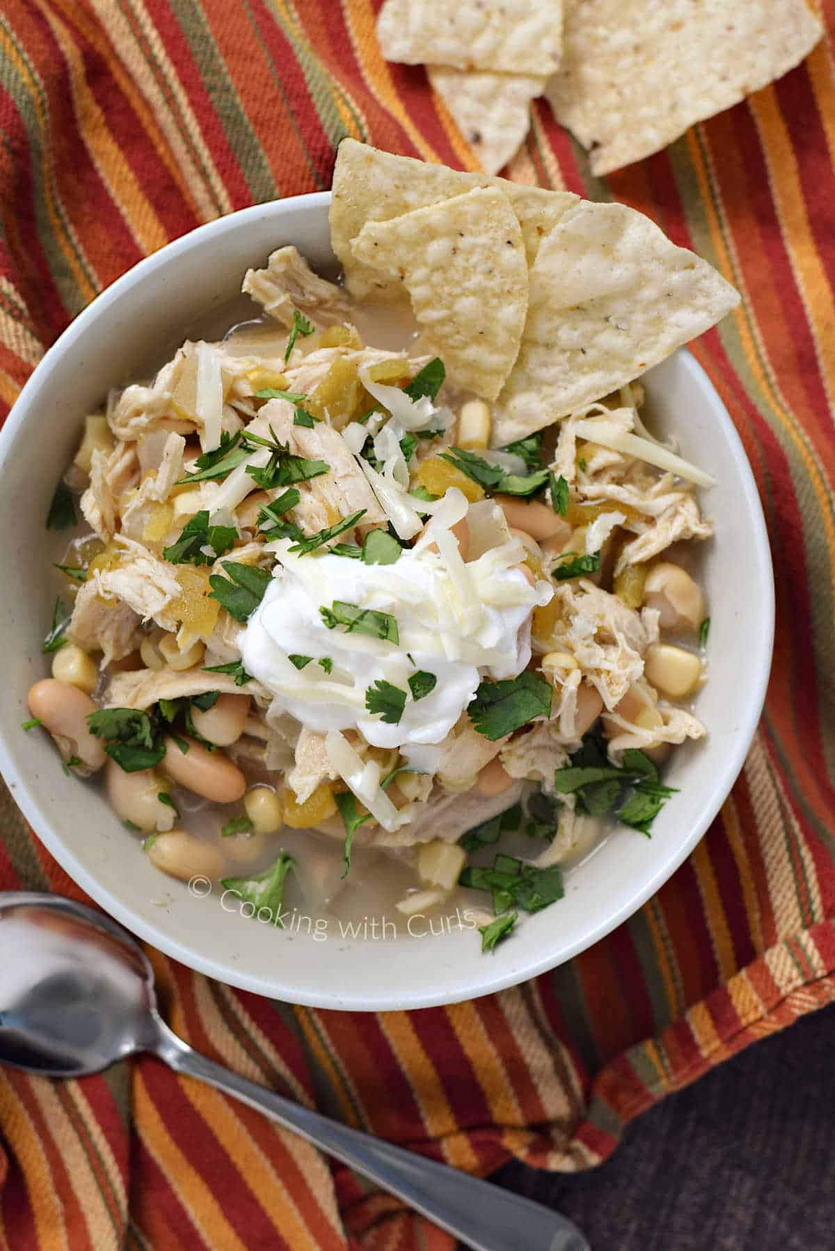 Looking down on a bowl of white bean chili with shredded chicken, corn, and green chilies topped with sour cream, cilantro, and tortilla chips.