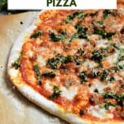 A whole pizza on parchment paper topped with tomato sauce, melted cheese, Italian sausage, and chopped kale with a wedge of parmesan in the background and title graphic across the top.