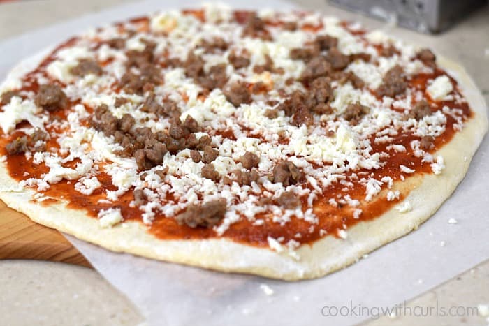 Pizza dough circle on a sheet of parchment paper topped with sauce, crumbled Italian sausage, and grated mozzarella cheese.