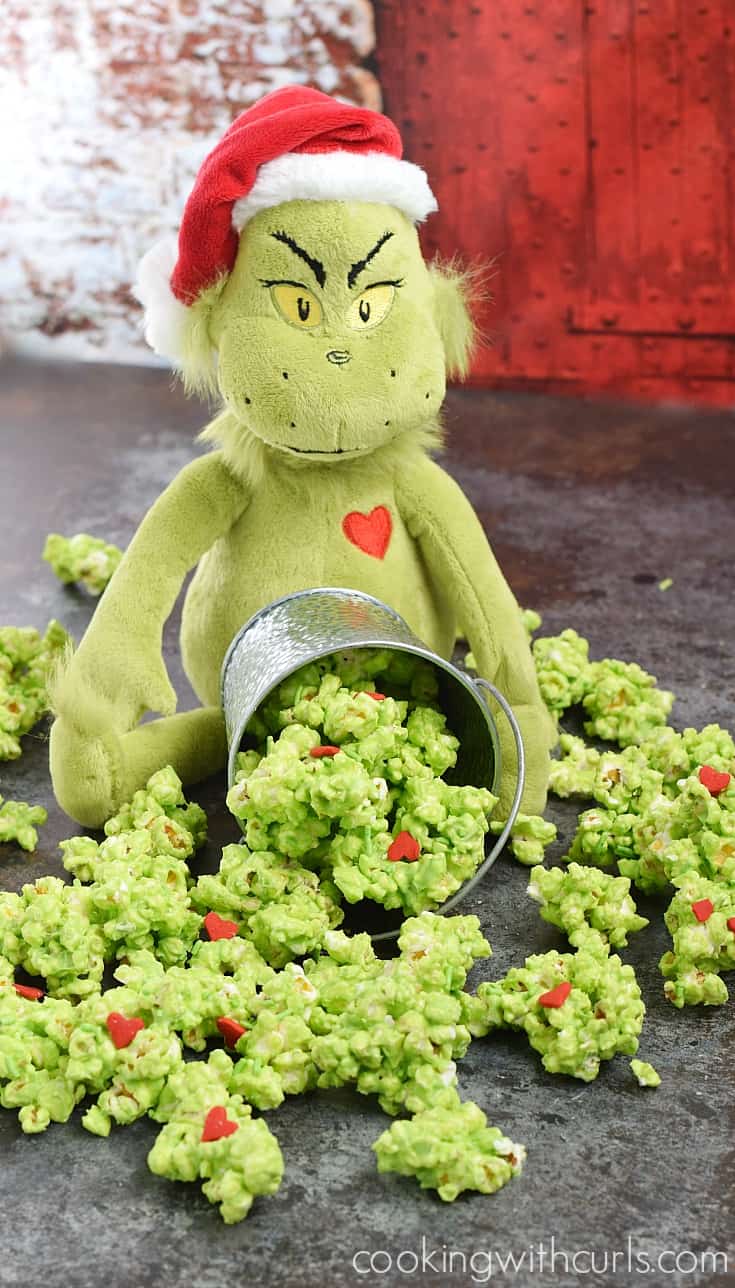A stuffed Grinch toy holding a silver bucket of spilled green popcorn with red heart and colored candies.
