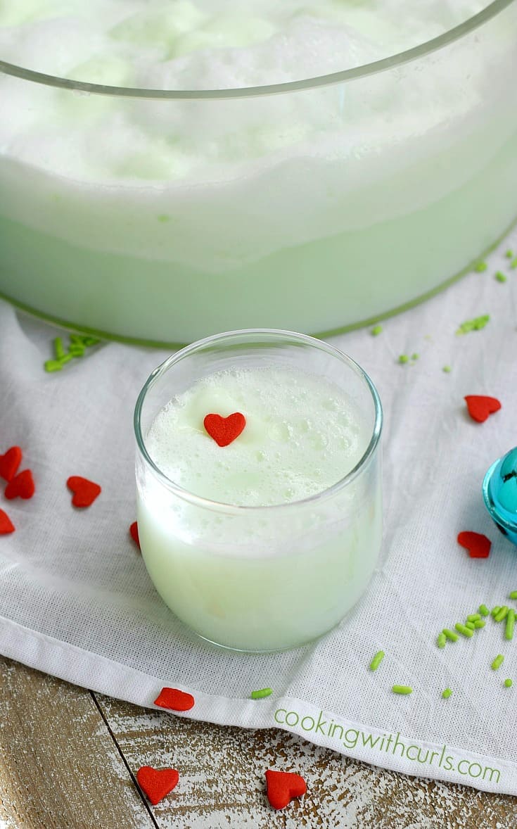 A glass of green Grinch punch in a glass with a red candy heart on top and a bowl of punch in the background.