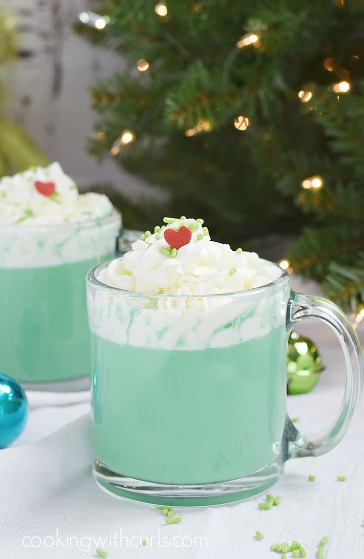 Two glass mugs of green hot chocolate topped with whipped cream, green sprinkles, and a red heart sitting in front of a lit Christmas tree.
