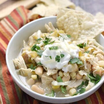 a bowl of white chicken chili topped with sour cream and green onions with tortilla chips stacked on the side