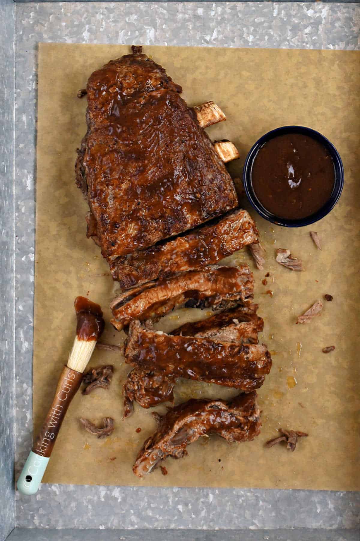A bbq sauce covered rack of pork ribs on a parchment paper lined metal tray with a bowl of sauce in the background.