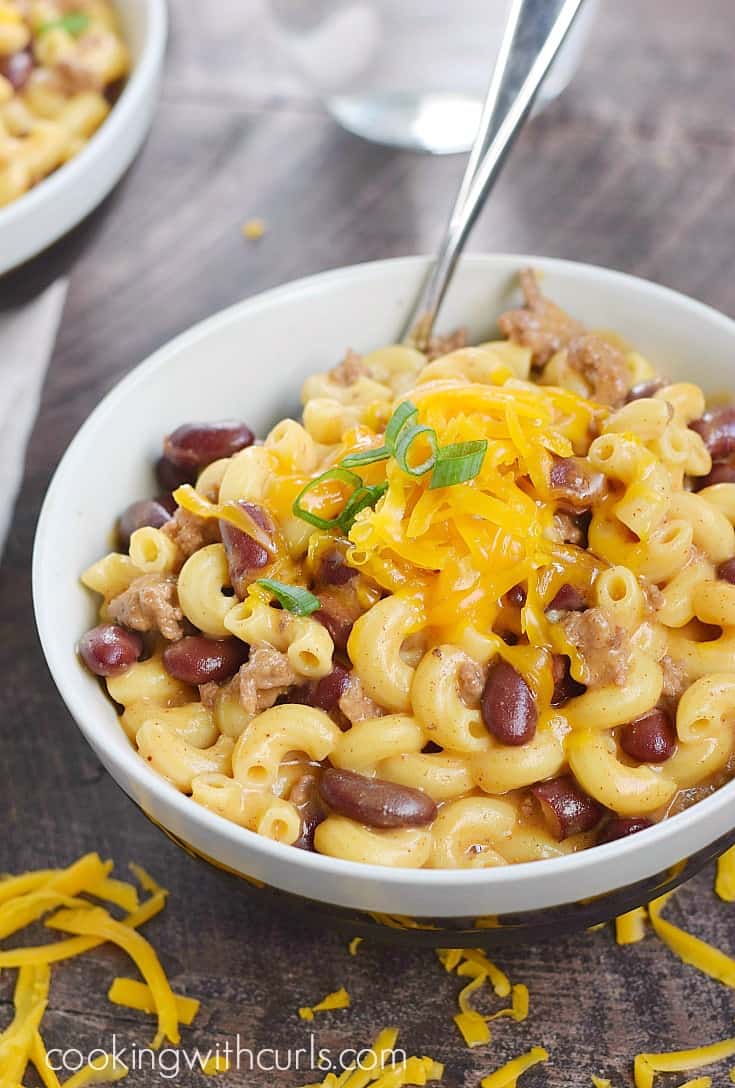 a big off-white bowl of macaroni, beans, and ground beef topped with shredded cheddar cheese