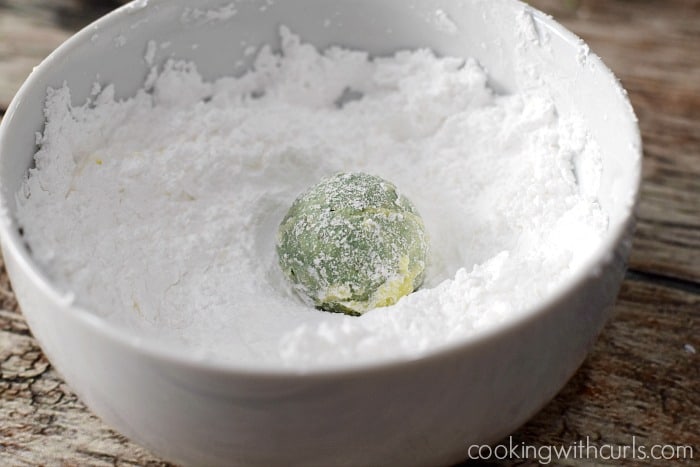 A green snowball cookie in a bowl filled with powdered sugar.