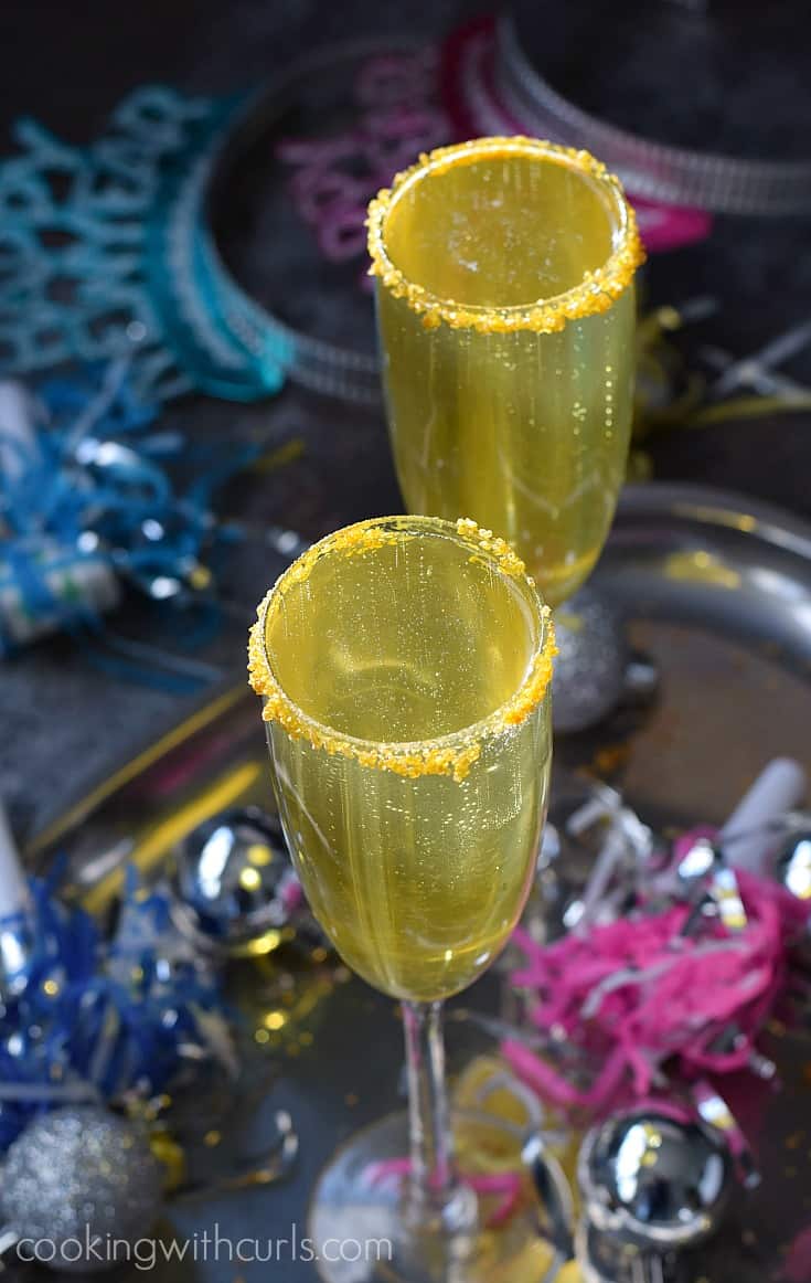 Two gold, bubbly cocktails in gold sugar rimmed champagne flutes surrounded by new year's eve hats.
