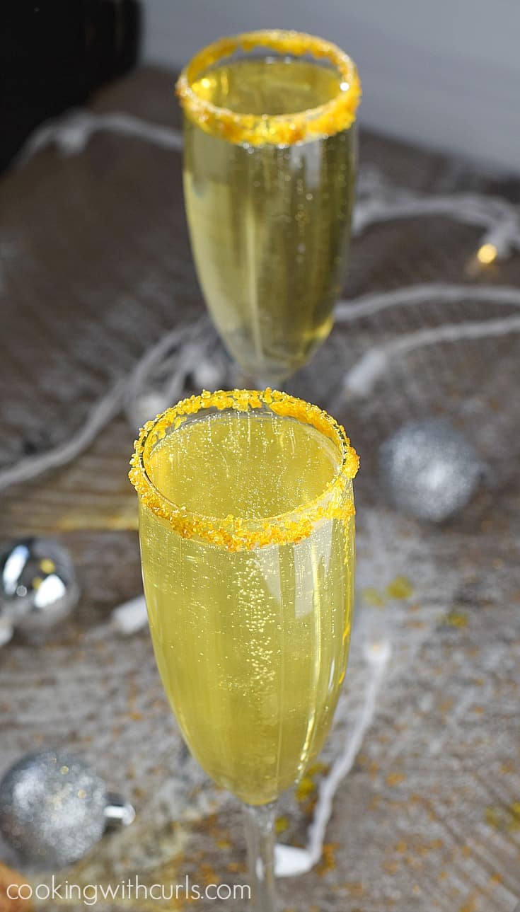 Two gold sugar rimmed champagne flutes filled with a bubbly gold cocktail surrounded by silver oranaments.