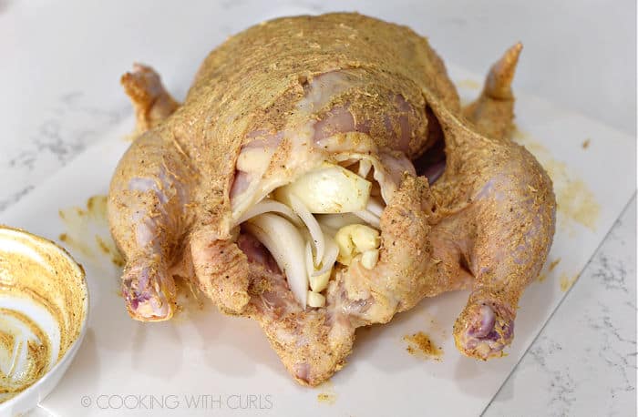 A butter rubbed whole chicken on a white cutting mat stuffed with onion slices and garlic cloves. 