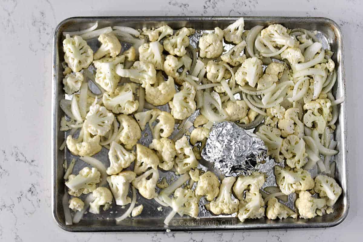 Seasoned chunks of cauliflower, onion slices, and foil wrapped garlic bulb on sheet pan.