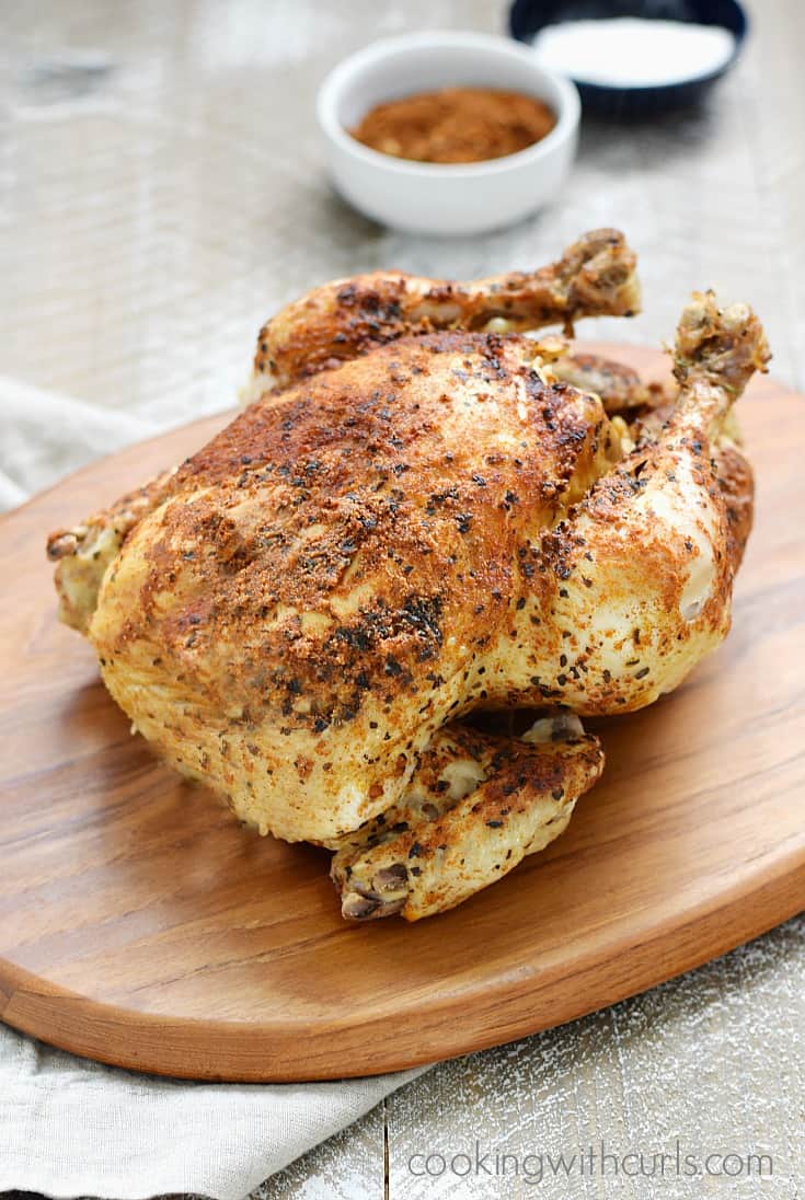 A whole, cooked chicken on a wood cutting board with small bowls of spices in the background.