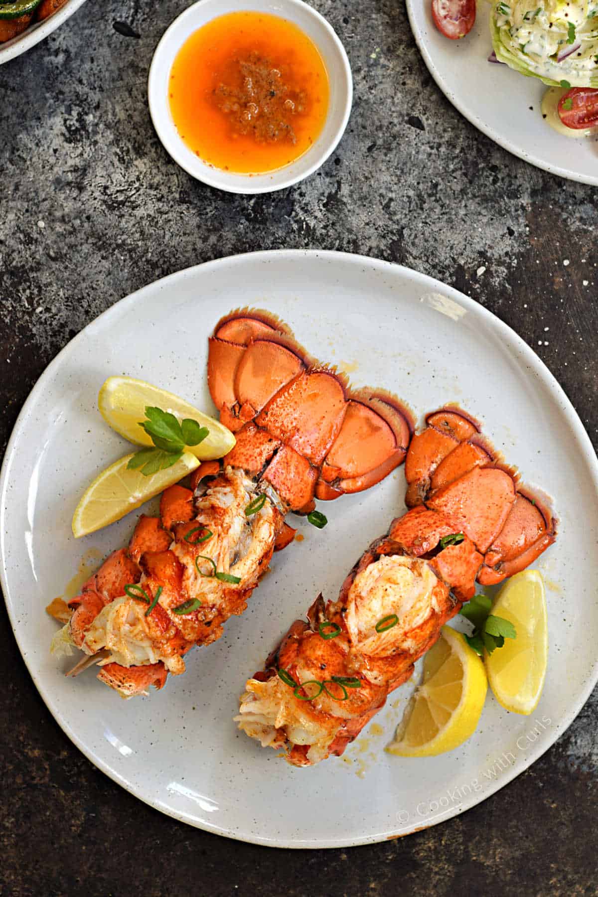 Two broiled lobster tails with lemon wedges and a bowl of garlic butter on the side.