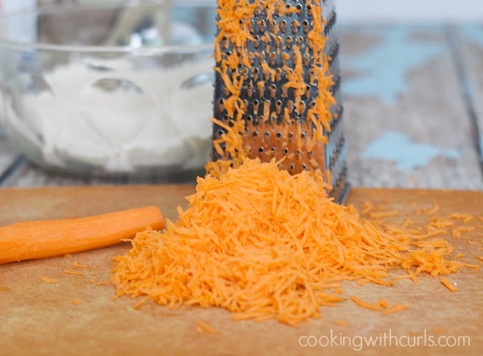 Grated carrots on a cutting board with a box grater and carrot in the background.