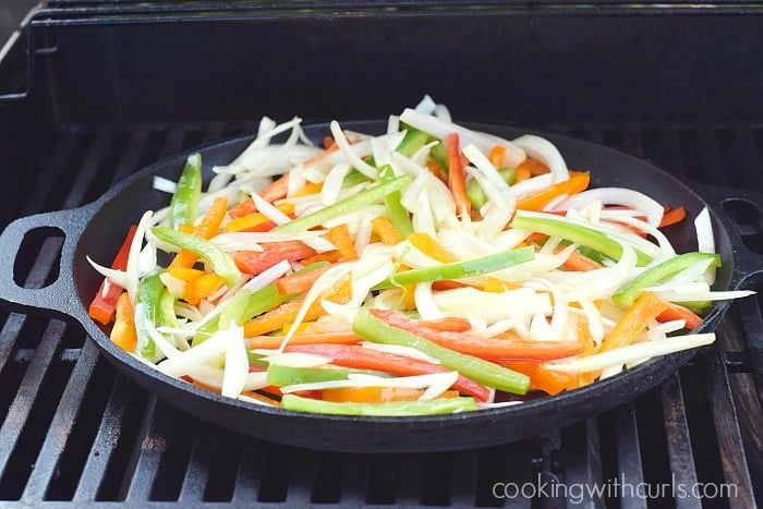 Thin strips of onions and bel peppers in a large cast iron skillet on a gas grill.