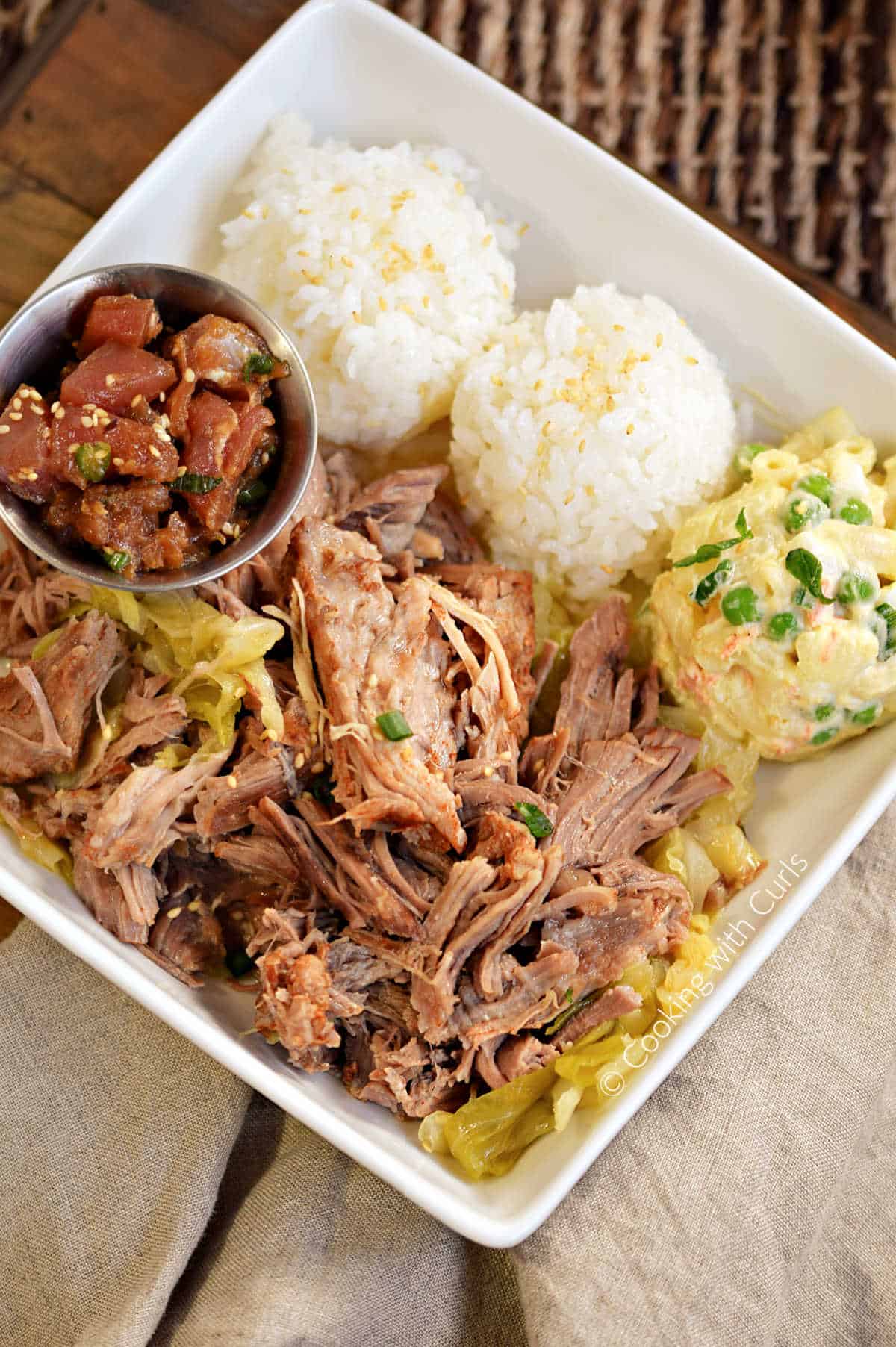 Looking down on a plate of shredded Kalua Pork surrounded by mounds of sticky rice, Hawaiian potato salad and poke. 