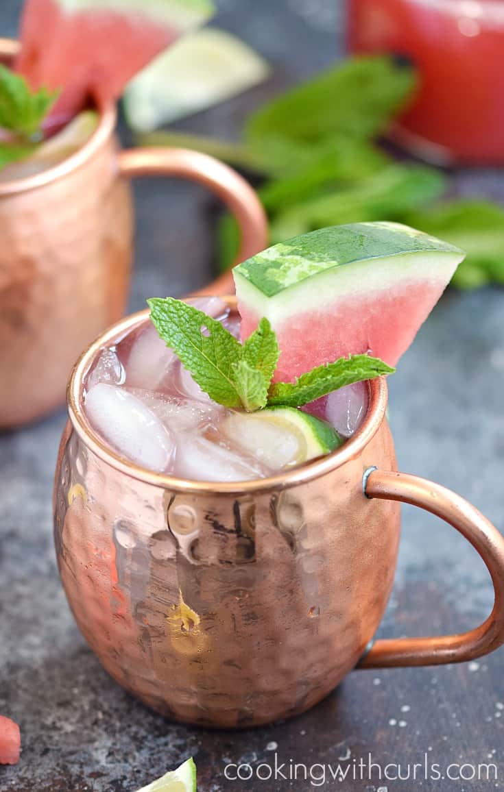 Close-up image of two copper mugs filled with pink liquid, lime wedge, mint sprig, and watermelon wedge.