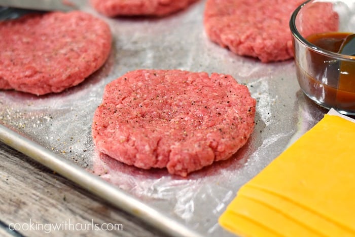 Four raw burger patties, a cup of barbecue sauce, and slices of cheddar cheese on a baking sheet.