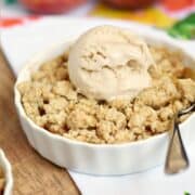 A scoop of ice cream on top of an oatmeal crumble over fresh peaches in a small white dish with fresh peaches in the background and title graphic across the top.