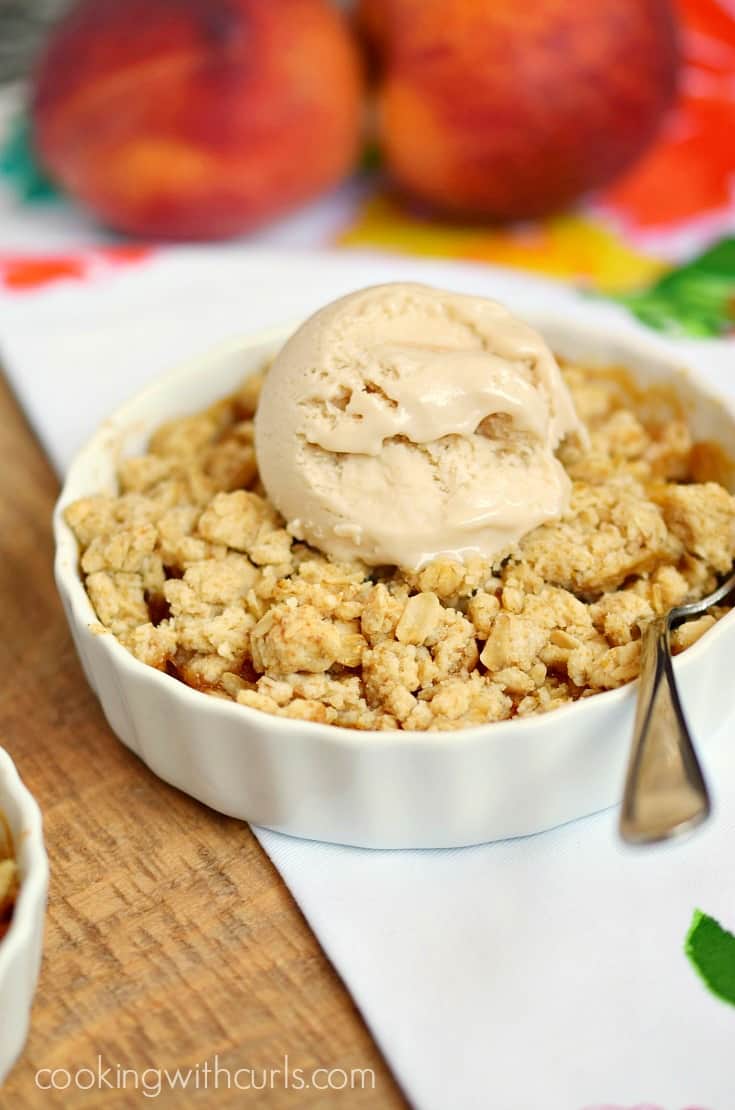 A scoop of ice cream on top of an oatmeal crumble over fresh peaches in a small white dish with fresh peaches in the background.
