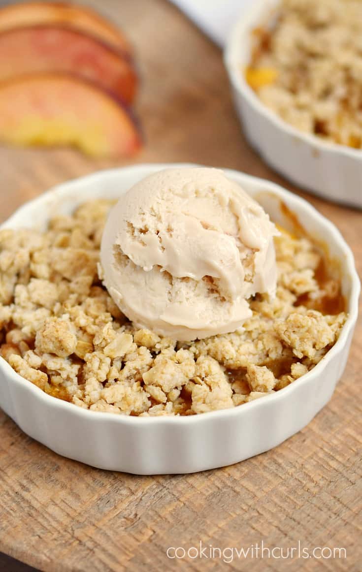 A scoop of ice cream on top of an oatmeal crumble over fresh peaches in a small white dish.