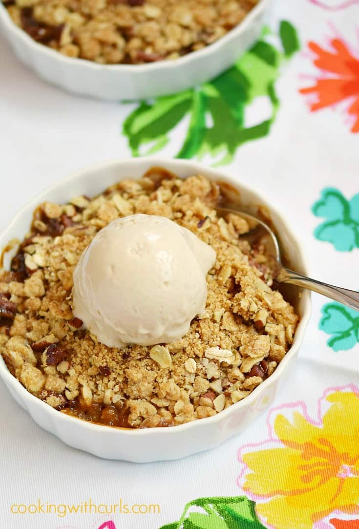 A scoop of ice cream on top of an oatmeal crumble over fresh peaches in a small white dish sitting on a floral napkin.