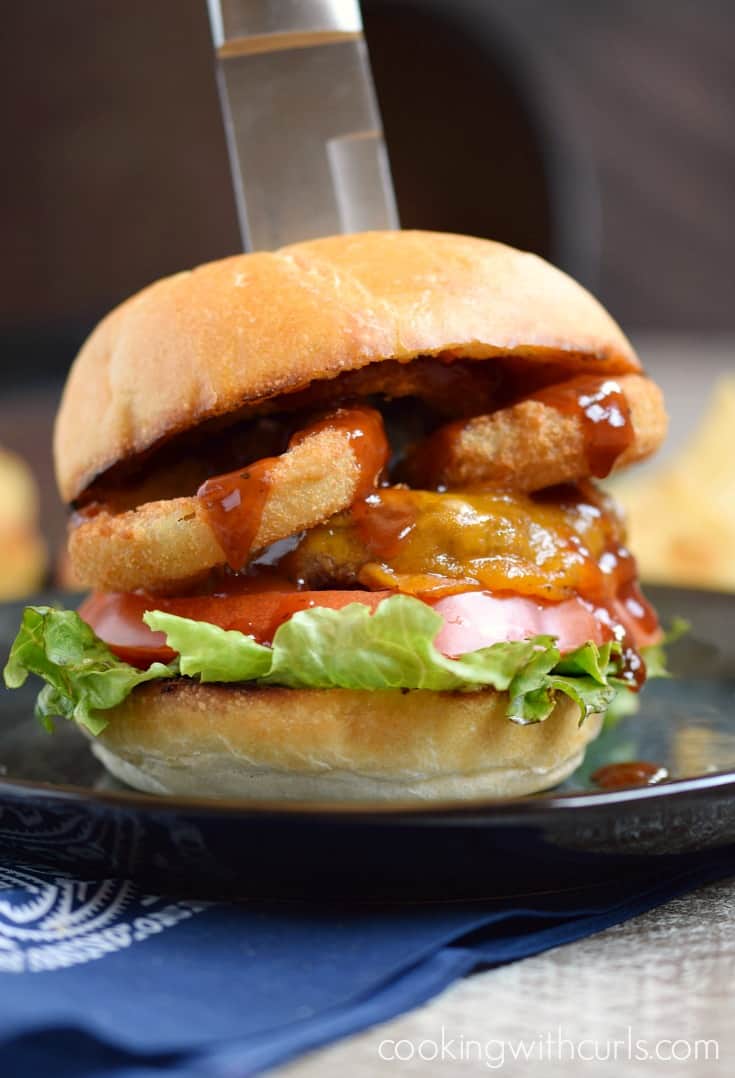 A cheeseburger topped with lettuce, tomato, onion rings, barbecue sauce and a toasted bun with a steak knife sticking out the top.