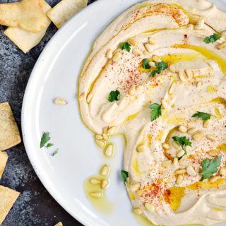 This thick and creamy Classic Homemade Hummus spread out across a large off-white plate topped with pine nuts, paprika and olive oil surrounded by pita chips