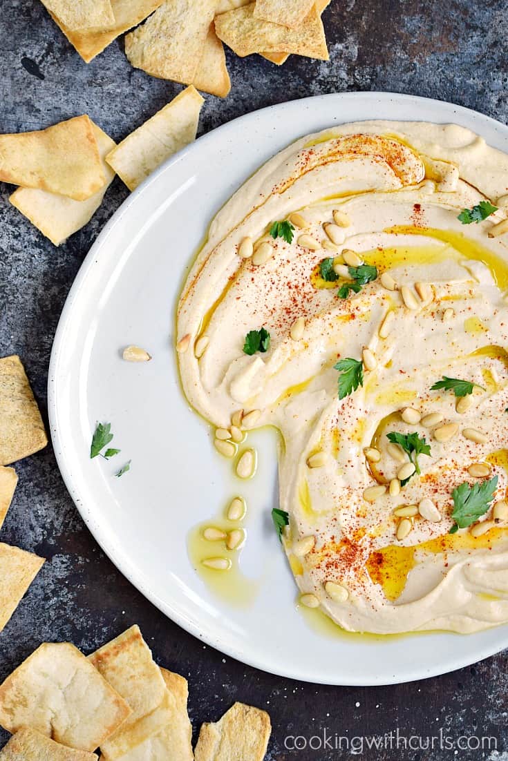 This thick and creamy Classic Homemade Hummus spread out across a large off-white plate topped with pine nuts, paprika and olive oil surrounded by pita chips