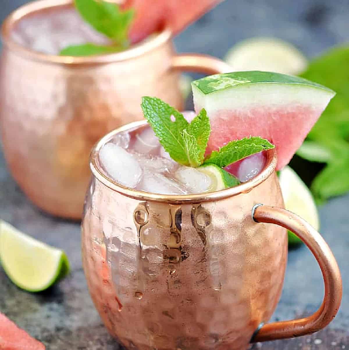 Two ice filled copper mugs with watermelon Moscow mule garnished with mint leaves and watermelon wedges.