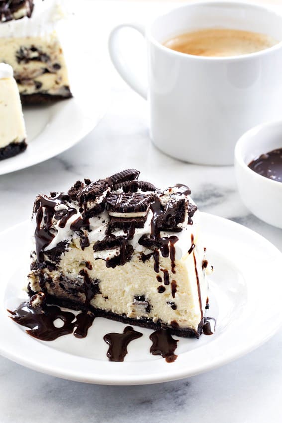 a slice of oreo cheesecake drizzled with chocolate sauce on a white plate with a cup of coffee and the whole cheesecake in the background.