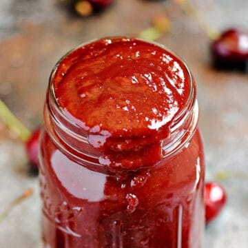 A glass jar overflowing with cherry barbecue sauce surrounded by fresh cherries.