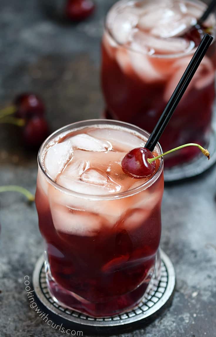 Two Cherry Whiskey Smash cocktails in clear glasses with a fresh cherry garnish and thin black straw.