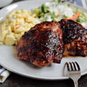 Two grilled bone-in chicken thighs with bbq sauce on a plate with potato salad and green salad.