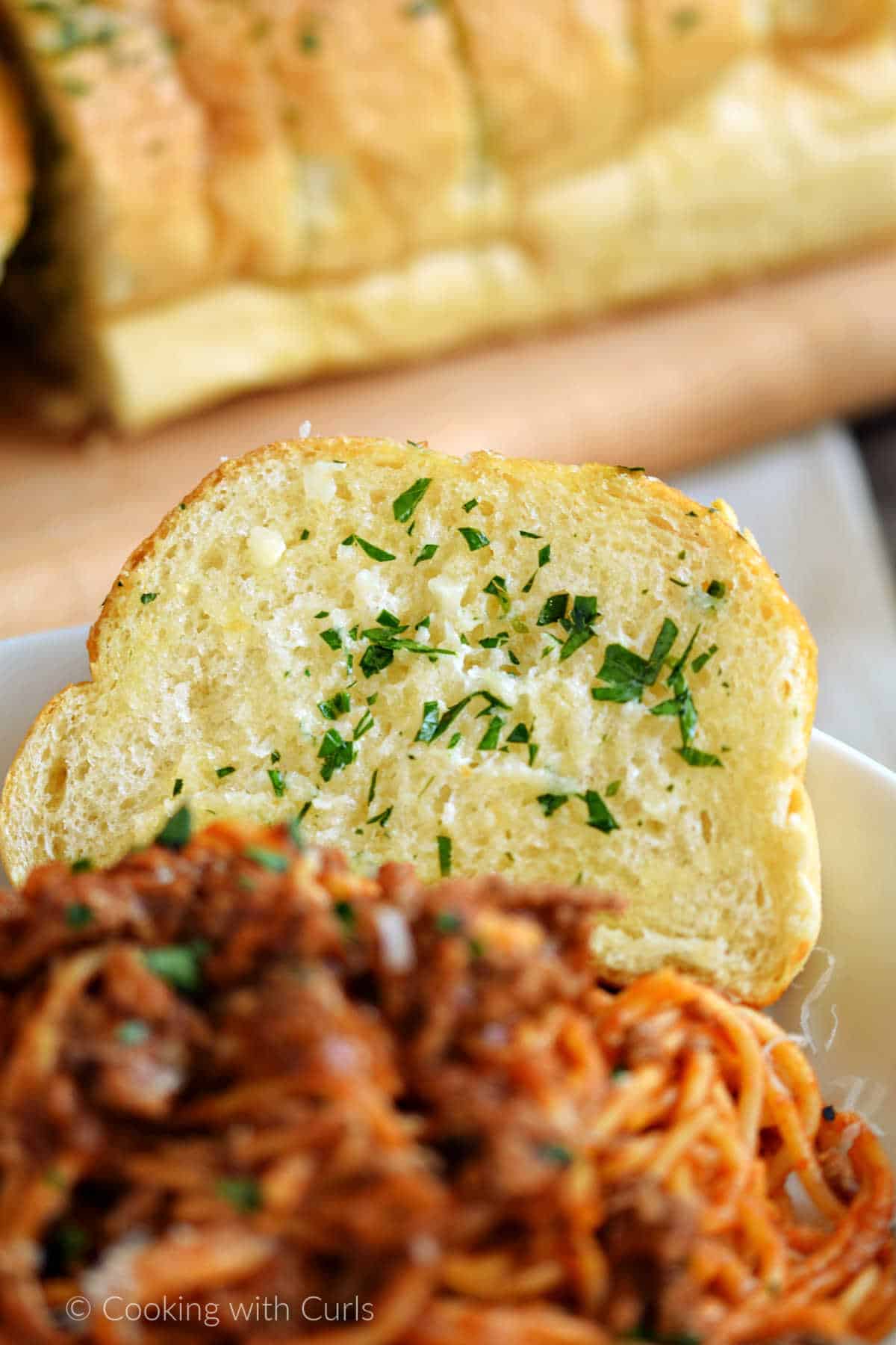 A slice of garlic bread on the edge of a bowl filled with spaghetti.