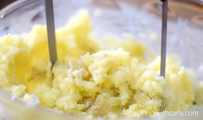Potatoes being mashed in a glass bowl with a masher tool.