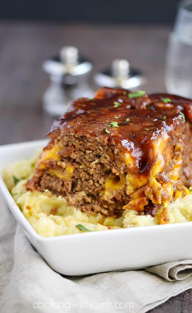 A loaf of Instant Pot Bacon Cheeseburger Meatloaf on a bed of creamy Mashed Potatoes in a square white bowl.