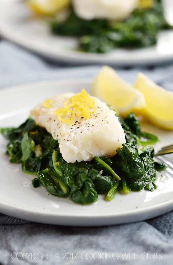 A cod filet on a bed of cooked spinach garnished with pepper and lemon zest and two lemon wedges on the side.