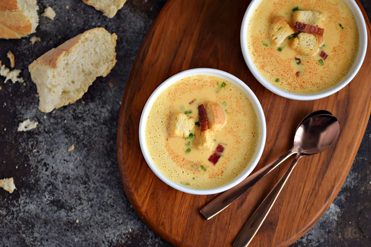 Two bowls of cheddar beer soup topped with croutons and bacon pieces sitting on a wooden serving board.