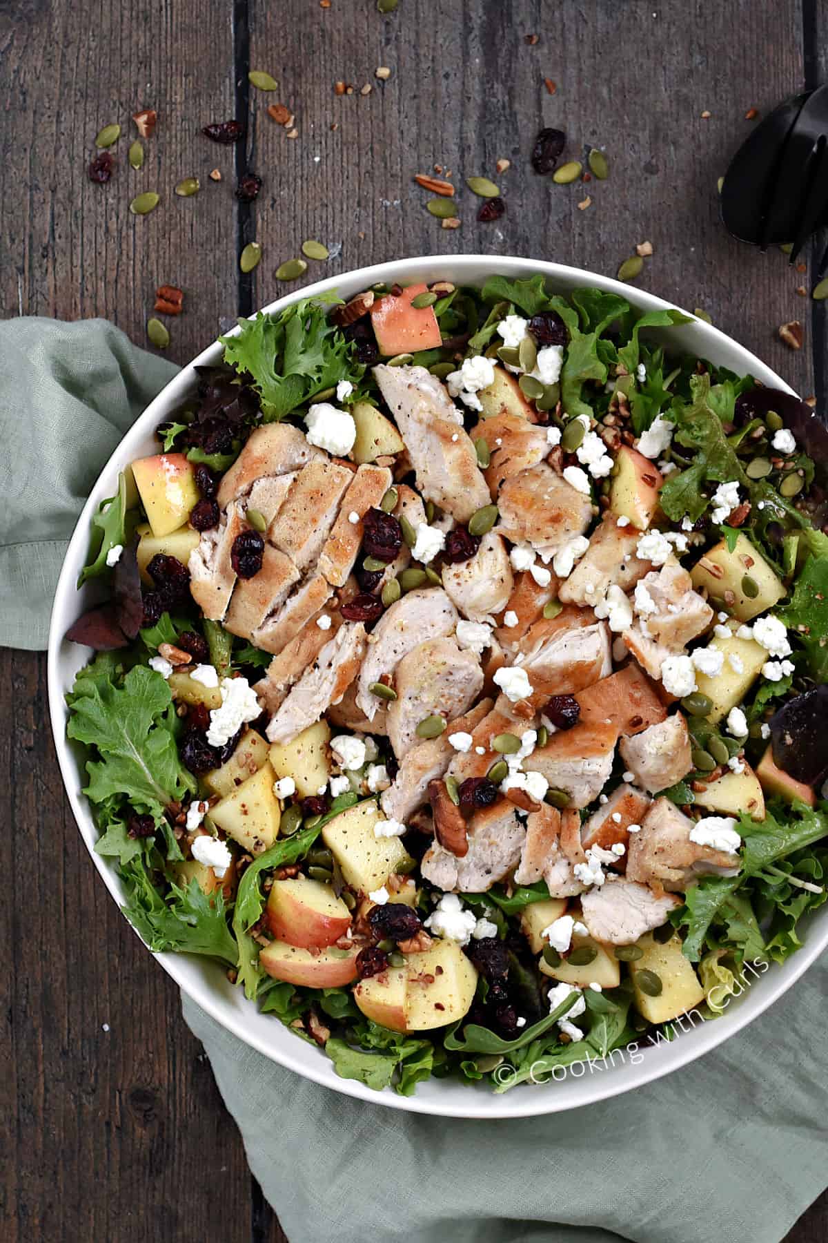 Looking down on a large bowl  filled with salad, apple chunks, dried cranberries, chicken, pepitas, and feta cheese. 