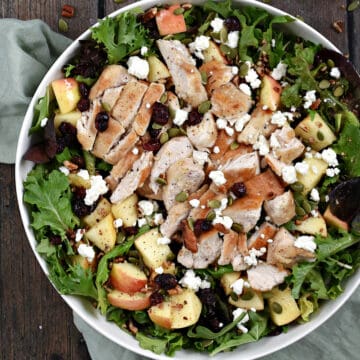 Looking down on a large bowl filled with salad, apple chunks, dried cranberries, chicken, pepitas, and feta cheese.