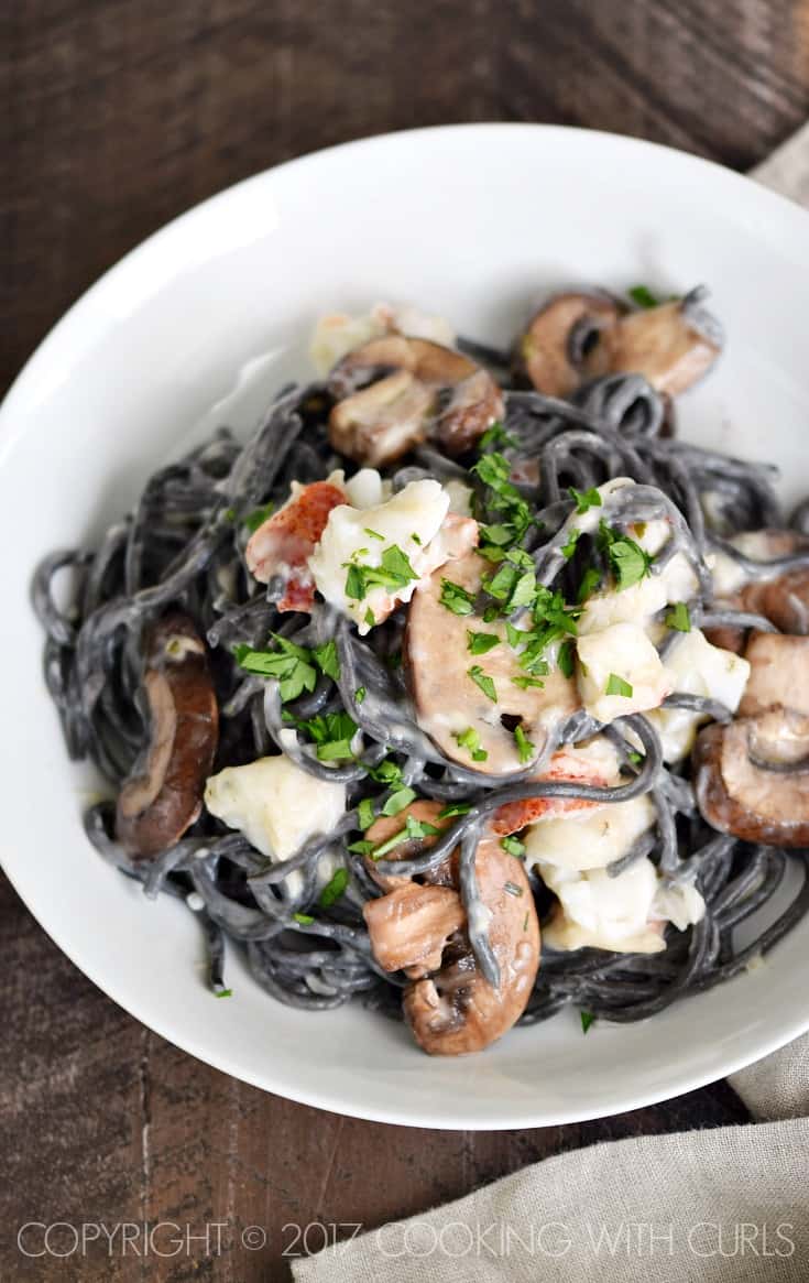 Looking down on a white bowl filled with black spaghetti pasta topped with a cream sauce, lobster chunks and sliced mushrooms.